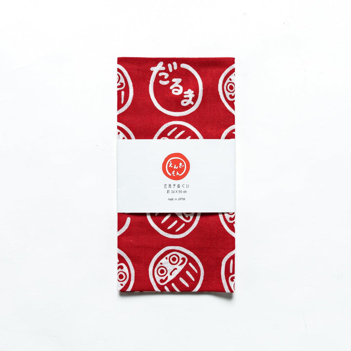 Folded red tenugui with white Daruma patterns, enclosed in a product sleeve, placed on a white background.