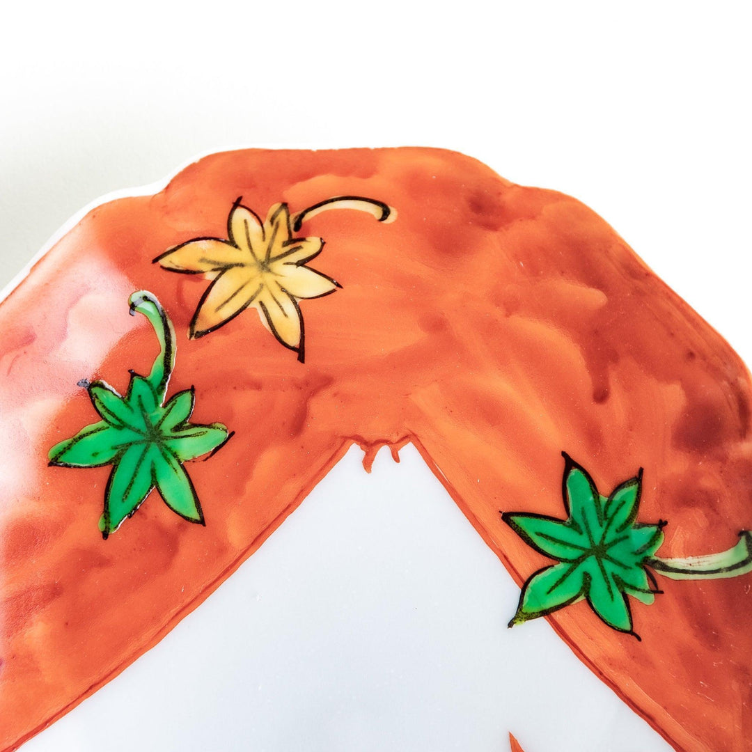 A white dish with a bright red border Mount Fuji, decorated with colorful maple leaves in red, yellow, and green.