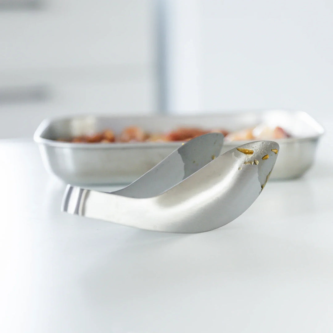 Japanese stainless steel kitchen tongs with curved tip design for easy grip and precise cooking, placed beside a metal tray