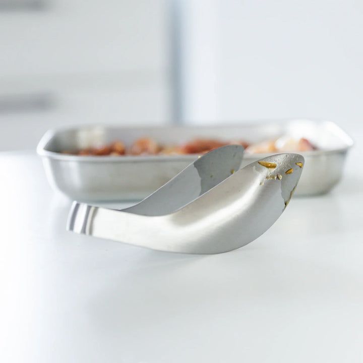 Japanese stainless steel kitchen tongs with curved tip design for easy grip and precise cooking, placed beside a metal tray