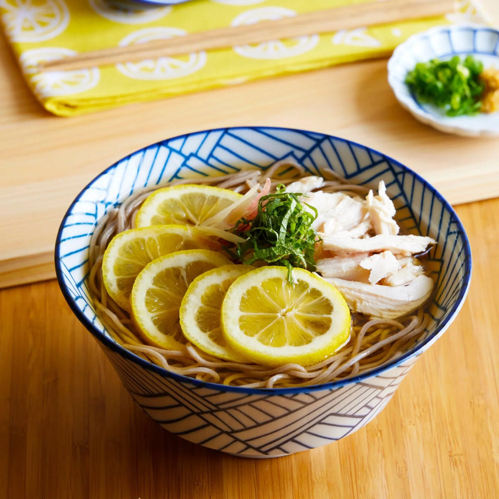 Blue and white ramen bowl with basket weave pattern filled with soba lemon chicken and herbs on table.