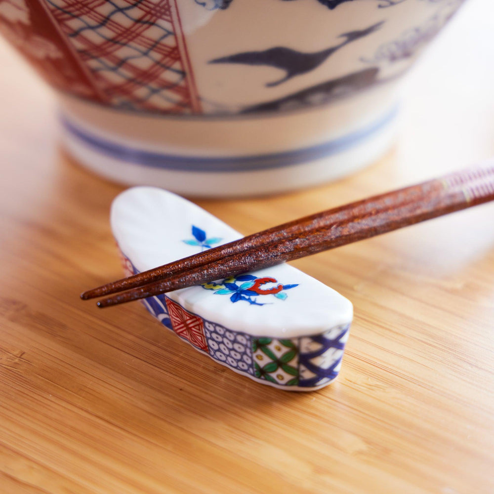 Japanese ceramic chopstick rest with colorful floral patterns, paired with wooden chopsticks and a ramen bowl in the back.