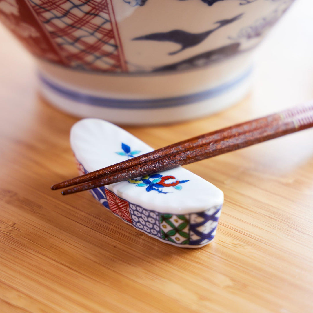 Japanese ceramic chopstick rest with colorful floral patterns, paired with wooden chopsticks and a ramen bowl in the back.
