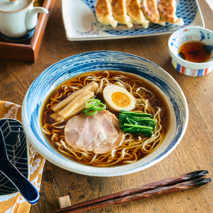 White ramen bowl with blue brushstroke pattern filled with noodles, topped with chashu, bamboo shoots, egg, and scallions.