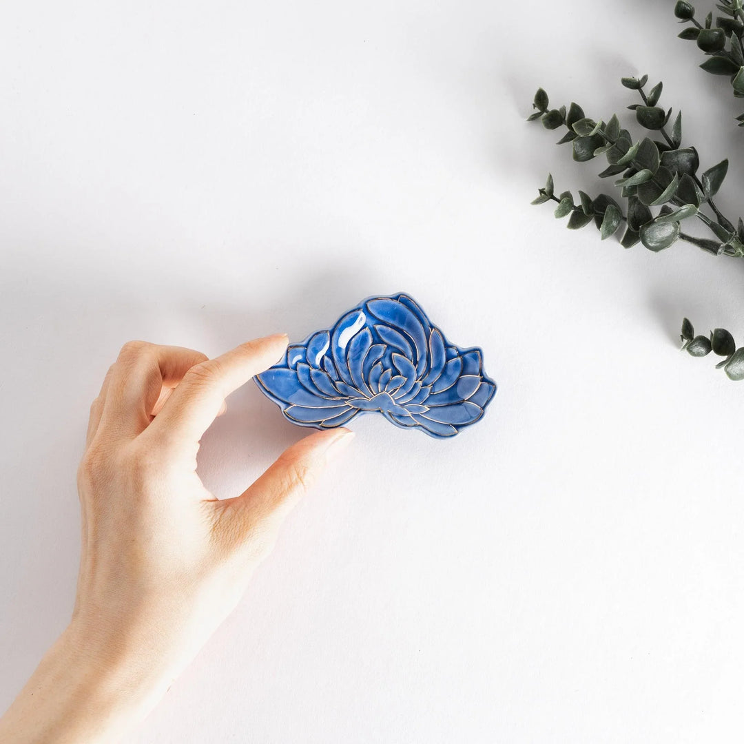 Hand holding blue mini Japanese sauce dish with gold chrysanthemum design and greenery in background.