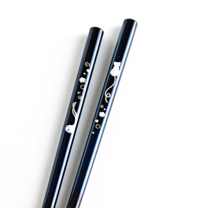 A pair of chopsticks, one blue and one red, each featuring a delicate white cat design near the top.