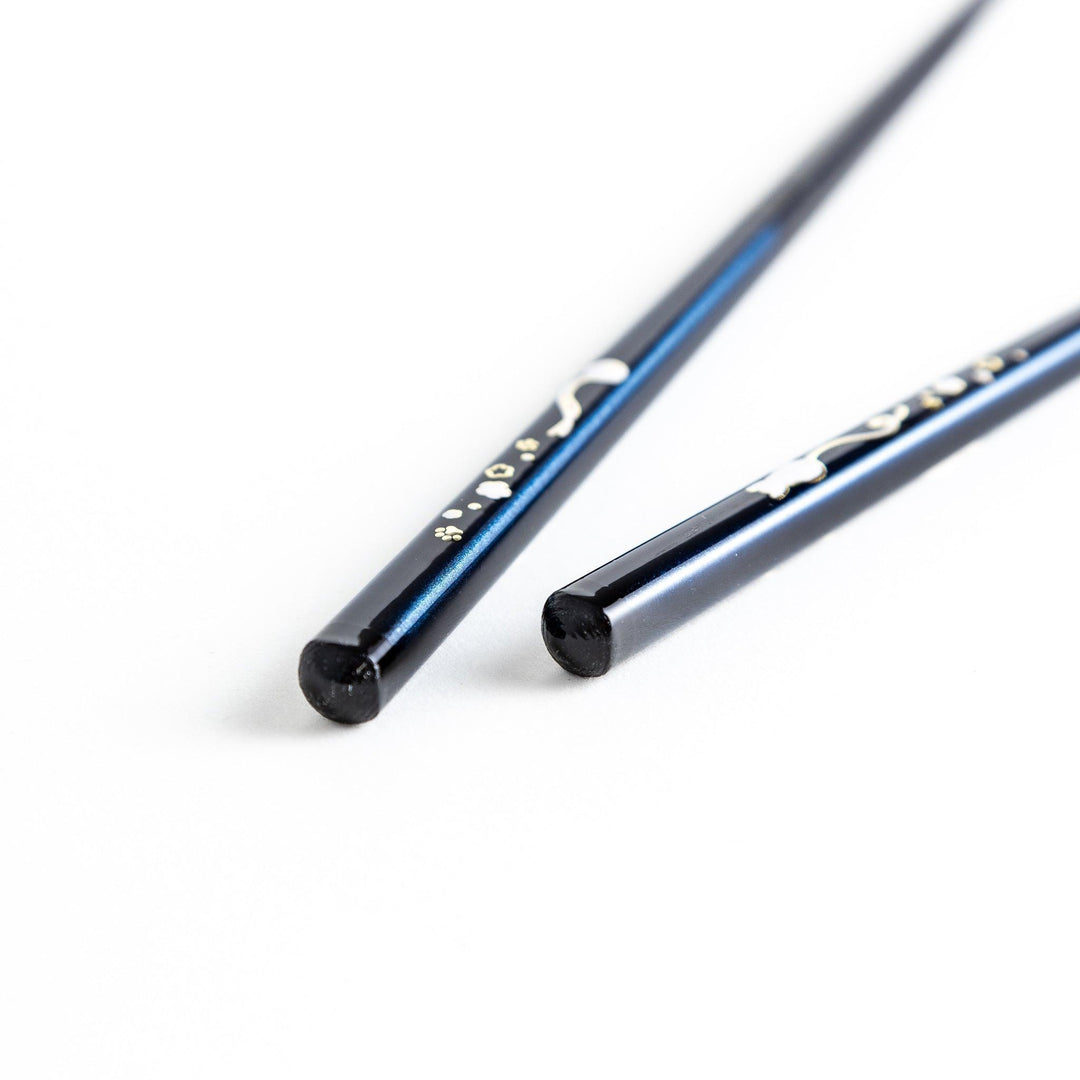 A pair of chopsticks, one blue and one red, each featuring a delicate white cat design near the top.