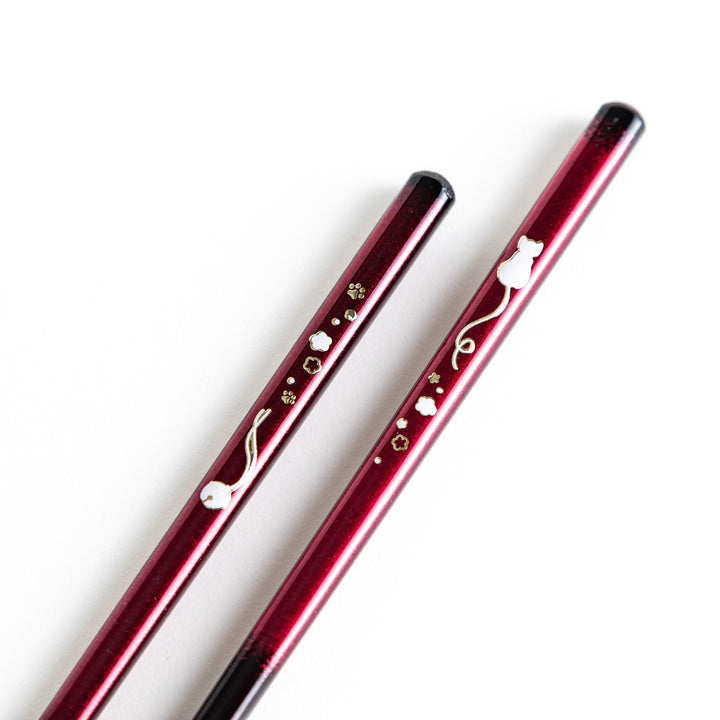 A pair of chopsticks, one blue and one red, each featuring a delicate white cat design near the top.