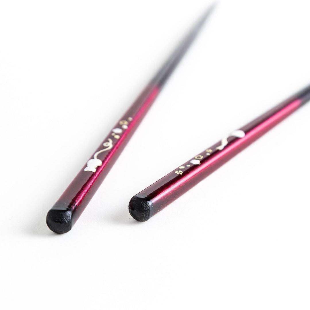 A pair of chopsticks, one blue and one red, each featuring a delicate white cat design near the top.