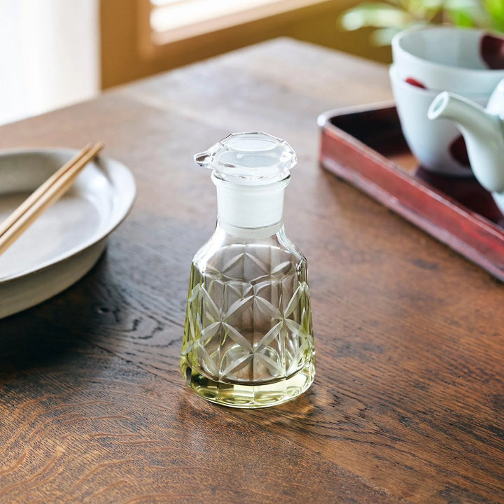 A glass soy sauce dispenser with a geometric cut pattern, filled with light-colored liquid on a wooden table.