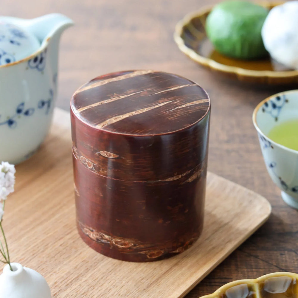 Dark brown cylindrical tea caddy made from natural cherry bark, placed on a wooden tray beside teacups and Japanese sweets.