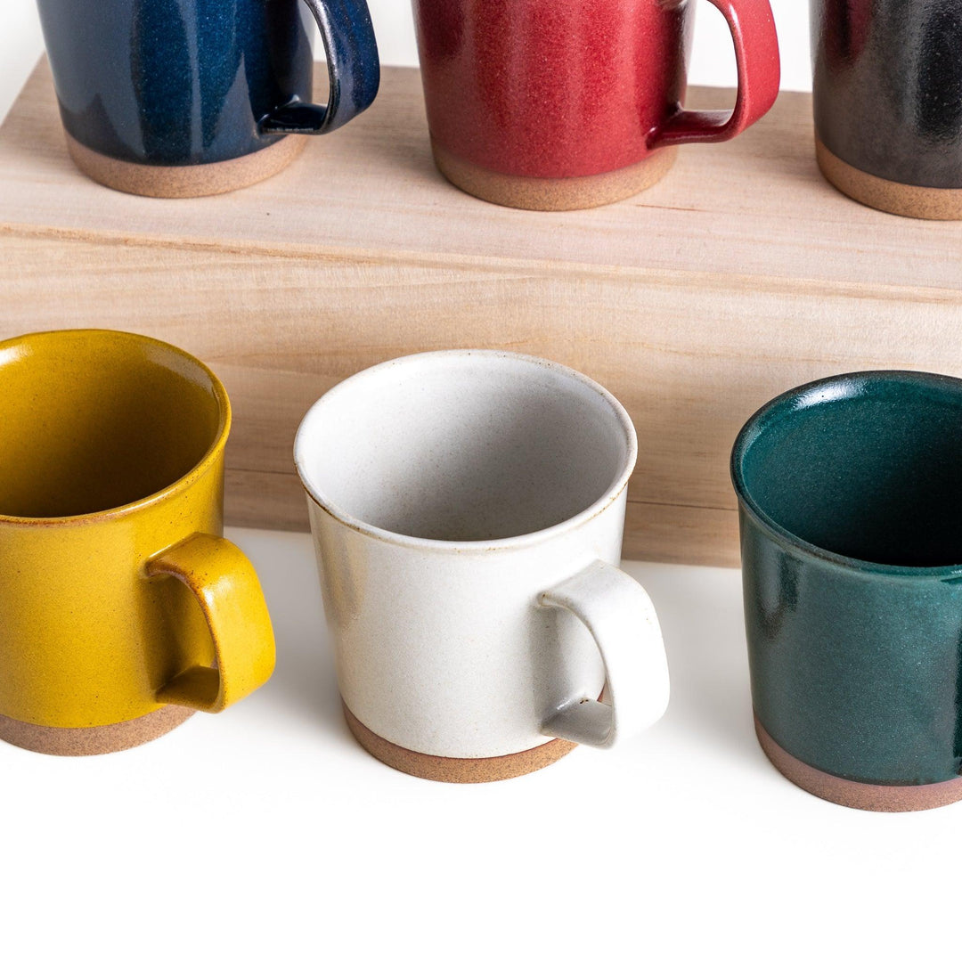 White Japanese mug with clay base beside colorful mugs in blue red yellow green and black on wood.