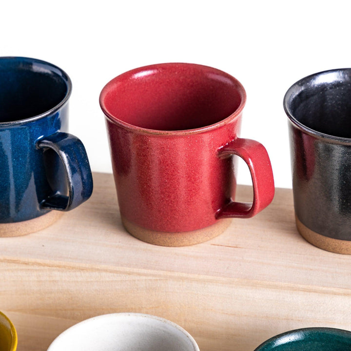 Colorful mug in blue red green yellow white or black with glossy finish and natural clay base on wooden block.