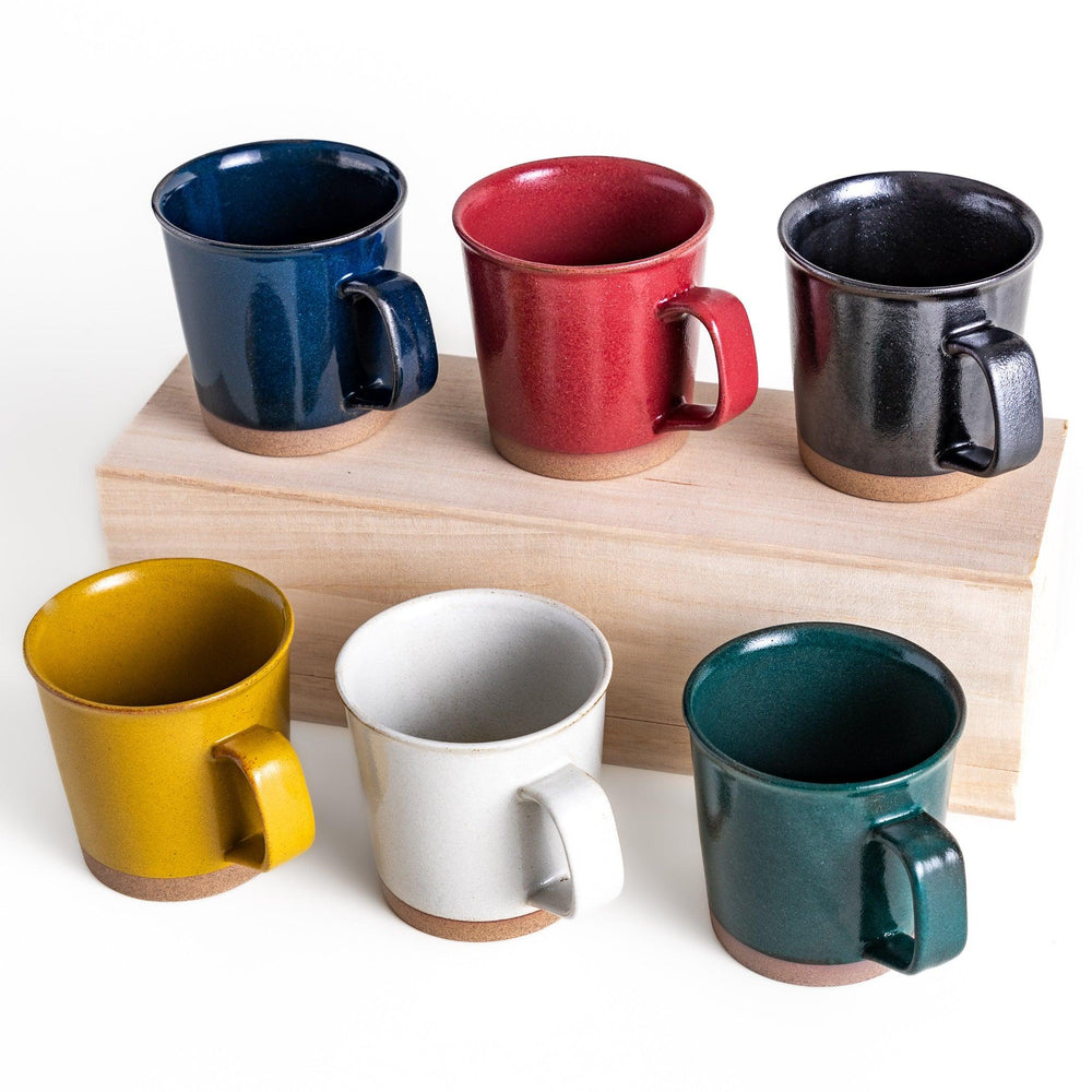 Colorful mug in blue red green yellow white or black with glossy finish and natural clay base on wooden block.