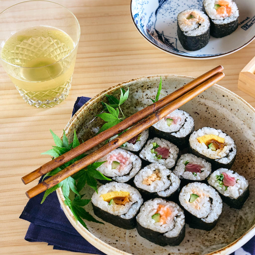 A pair of serving chopsticks resting on a ceramic bowl filled with assorted sushi rolls.