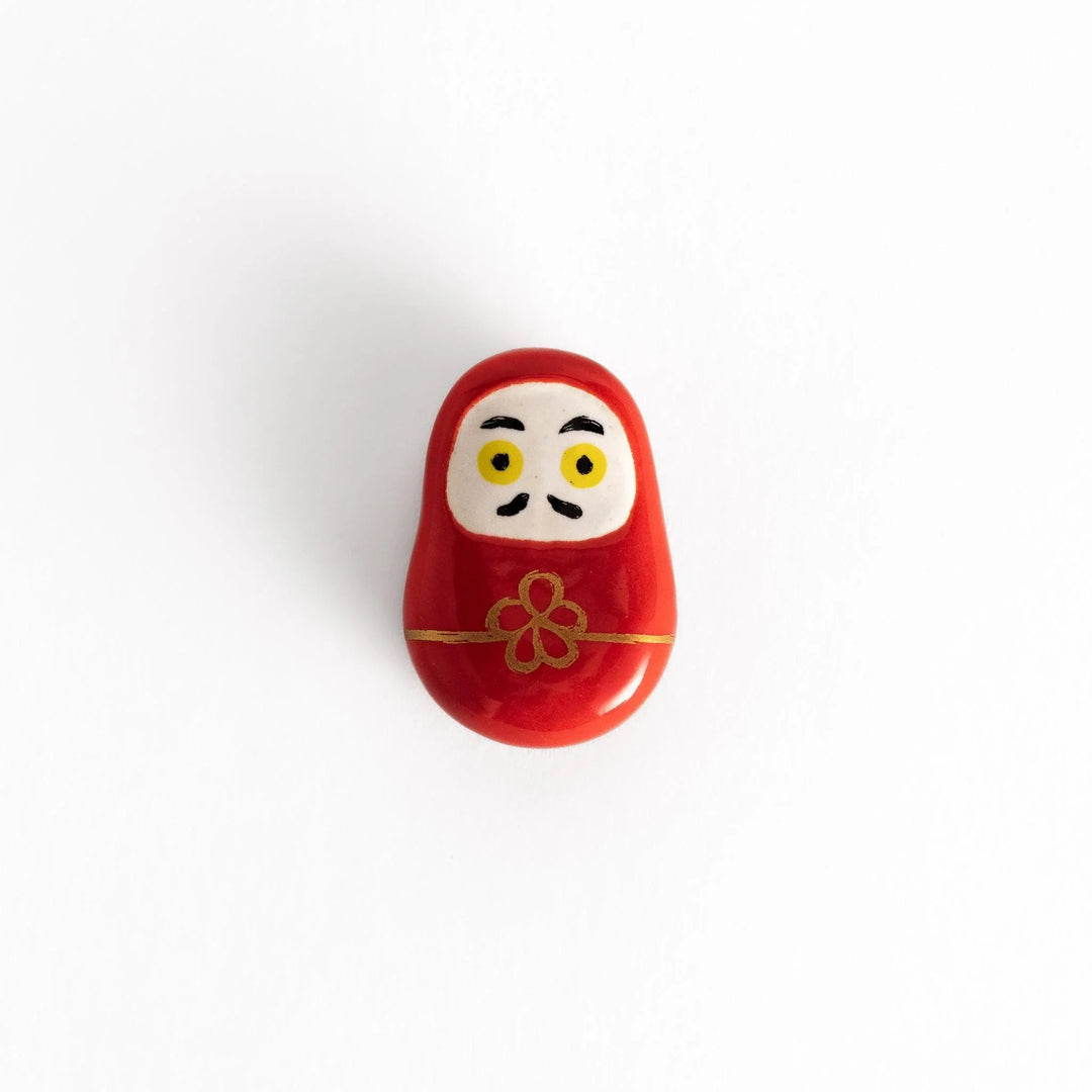 Red daruma doll chopstick rest with gold cord and flower motif symbolizing luck in Japanese dining.