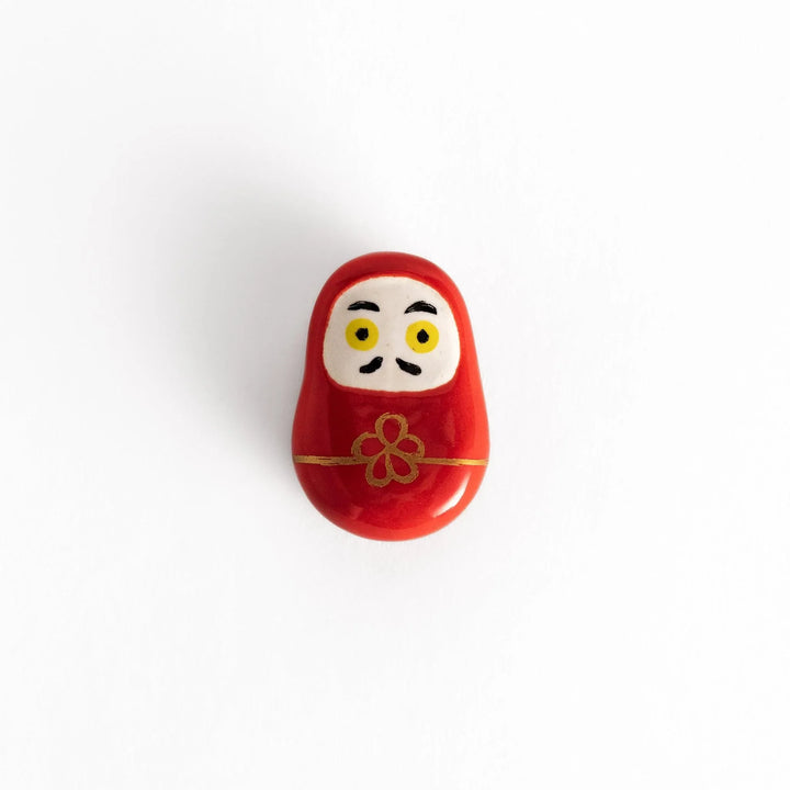 Red daruma doll chopstick rest with gold cord and flower motif symbolizing luck in Japanese dining.