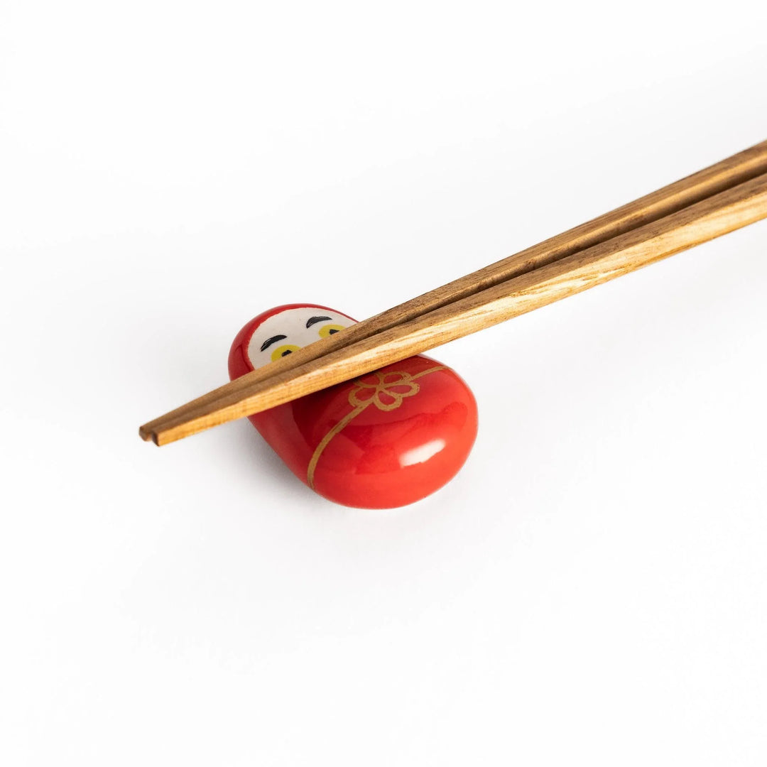 Red daruma doll chopstick rest with gold cord and flower motif symbolizing luck in Japanese dining.