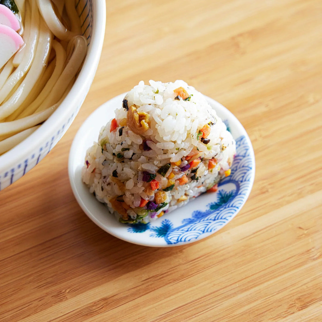 A small white plate with a blue wave pattern, holding a colorful mixed vegetable onigiri made with seasoned rice and chopped ingredients.