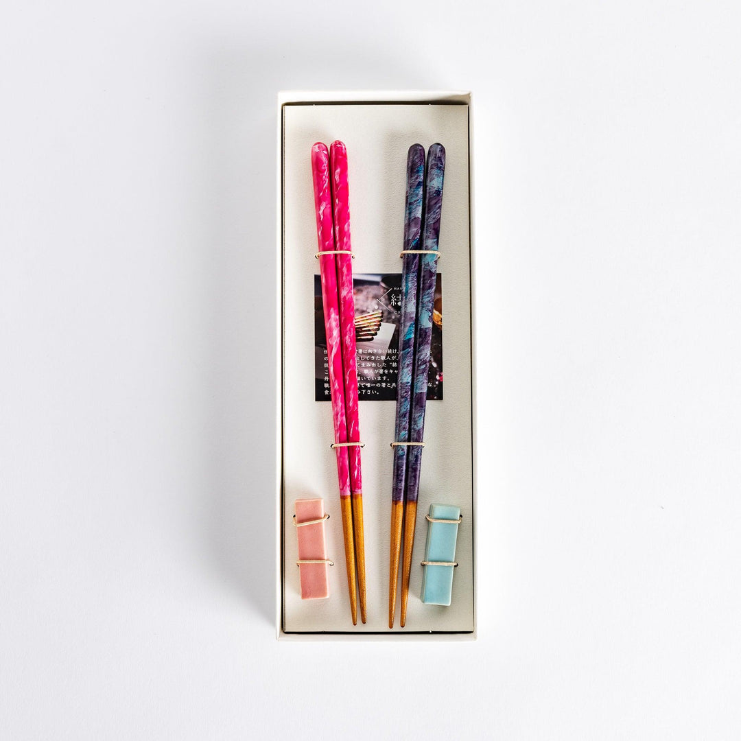 Two pairs of chopsticks one pink with swirls and one dark with blue accents tied together in box.