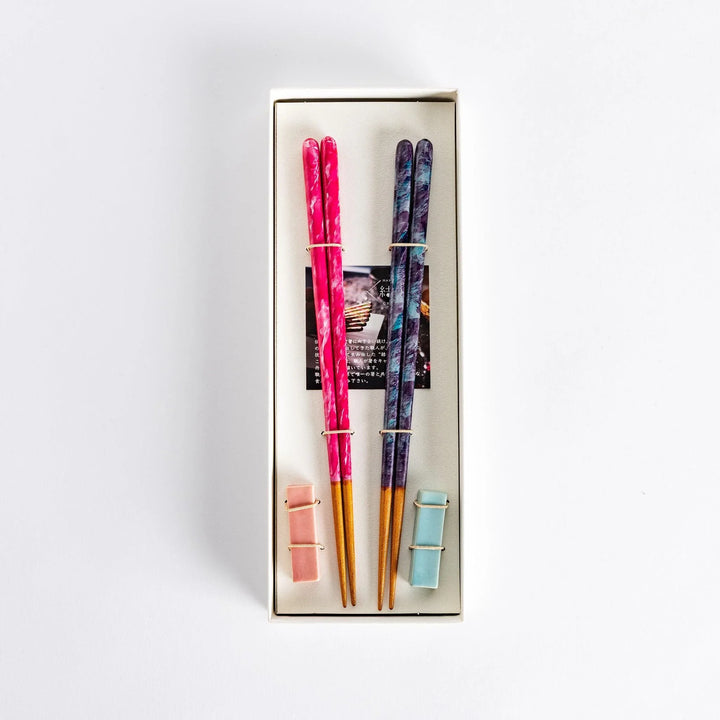 Two pairs of chopsticks one pink with swirls and one dark with blue accents tied together in box.