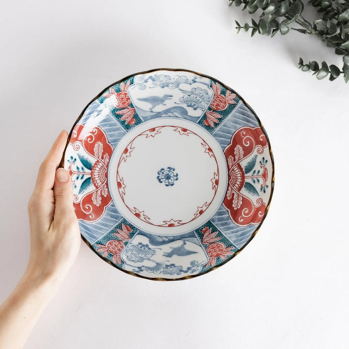A hand holding the decorative plate, showcasing the detailed artwork in red, blue, and green against a white background.