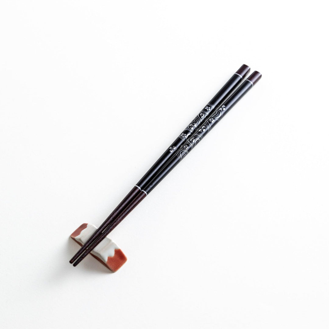Two pairs of chopsticks, one black and one red, each adorned with intricate floral patterns on a dark background.