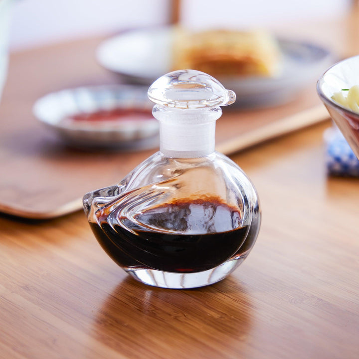 Handcrafted glass soy sauce dispenser with a swirl design, placed on a wooden table during a Japanese meal setting.