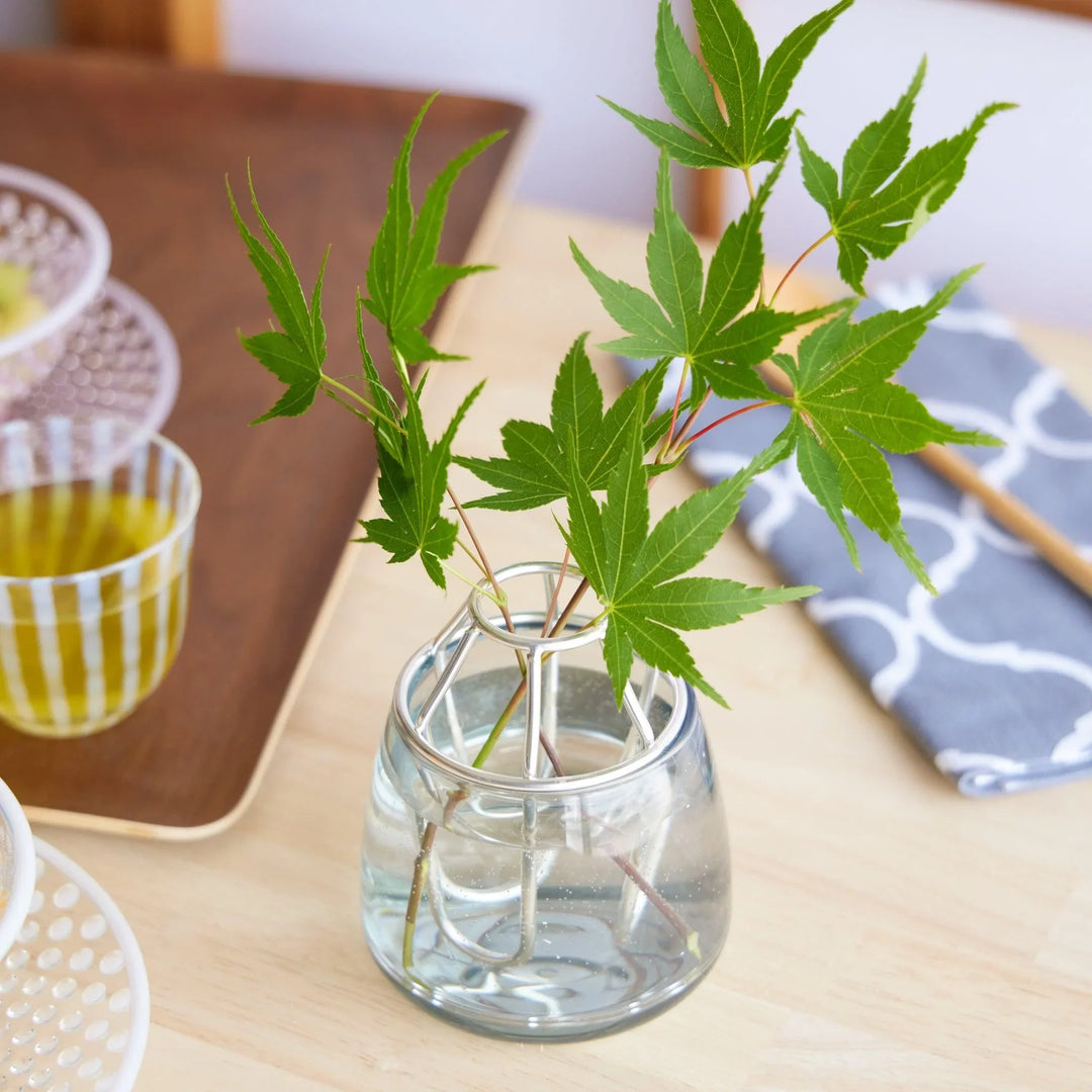 Clear glass vase with a silver flower holder, displaying fresh green maple leaves on a wooden table with summer tableware.
