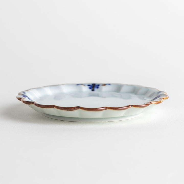Round white porcelain sauce dish with scalloped edges, adorned with a simple blue floral pattern and brown accent trim.