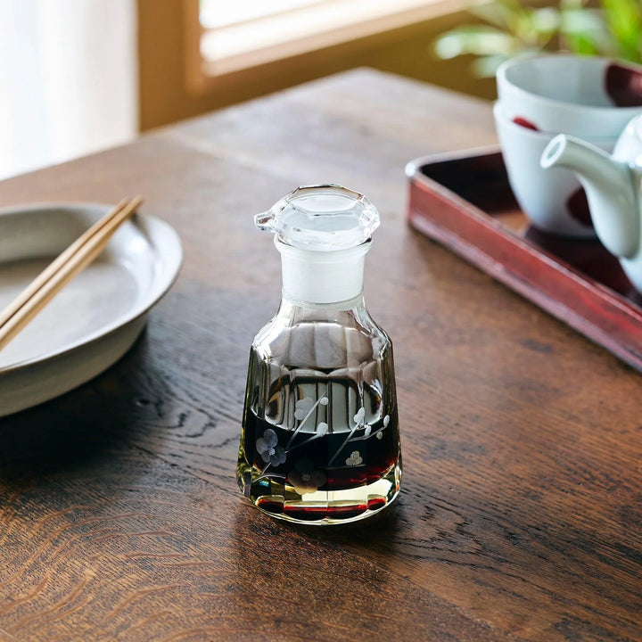 Glass soy sauce dispenser with etched floral pattern, partially filled with dark soy sauce, on a wooden table.