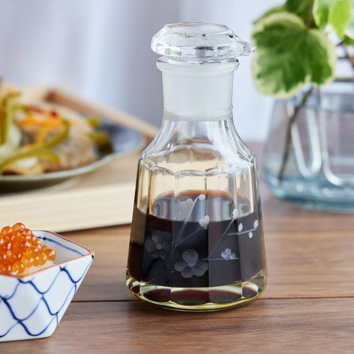 A clear glass soy sauce dispenser with etched plum blossom motifs, placed on a wooden table with Japanese dishes.