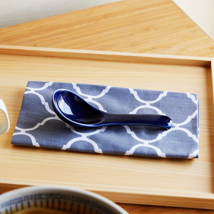 A blue ceramic spoon resting on a white and blue patterned tenugui, placed on a wooden tray.