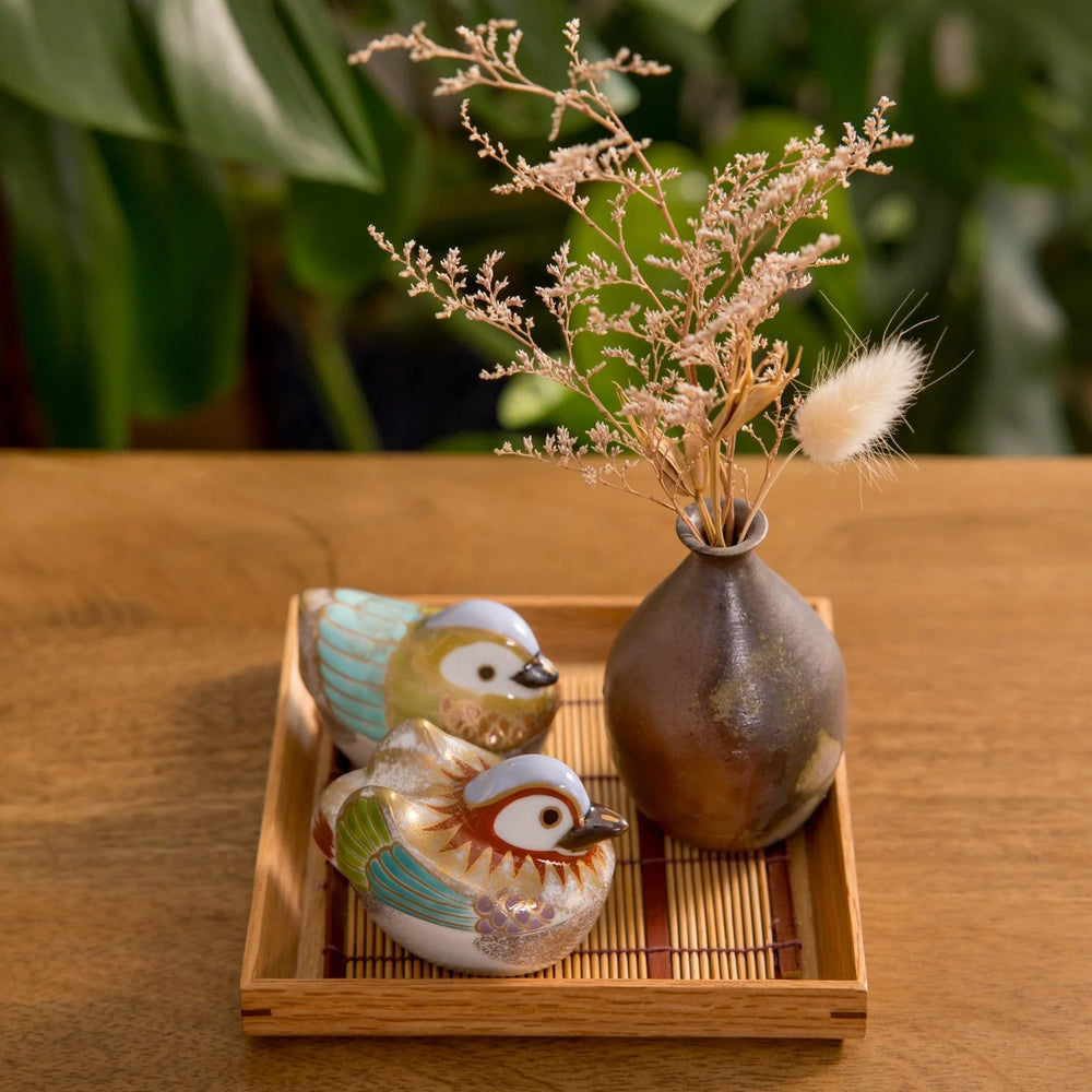 Two colorful bird shaped bowls on bamboo mat tray beside vase with dried flowers for cozy table decor.