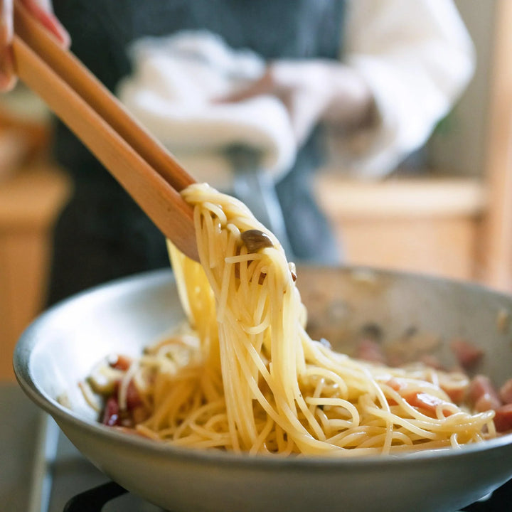 Wooden tongs lifting cooked spaghetti from a frying pan with visible ingredients like mushrooms and bacon in a kitchen