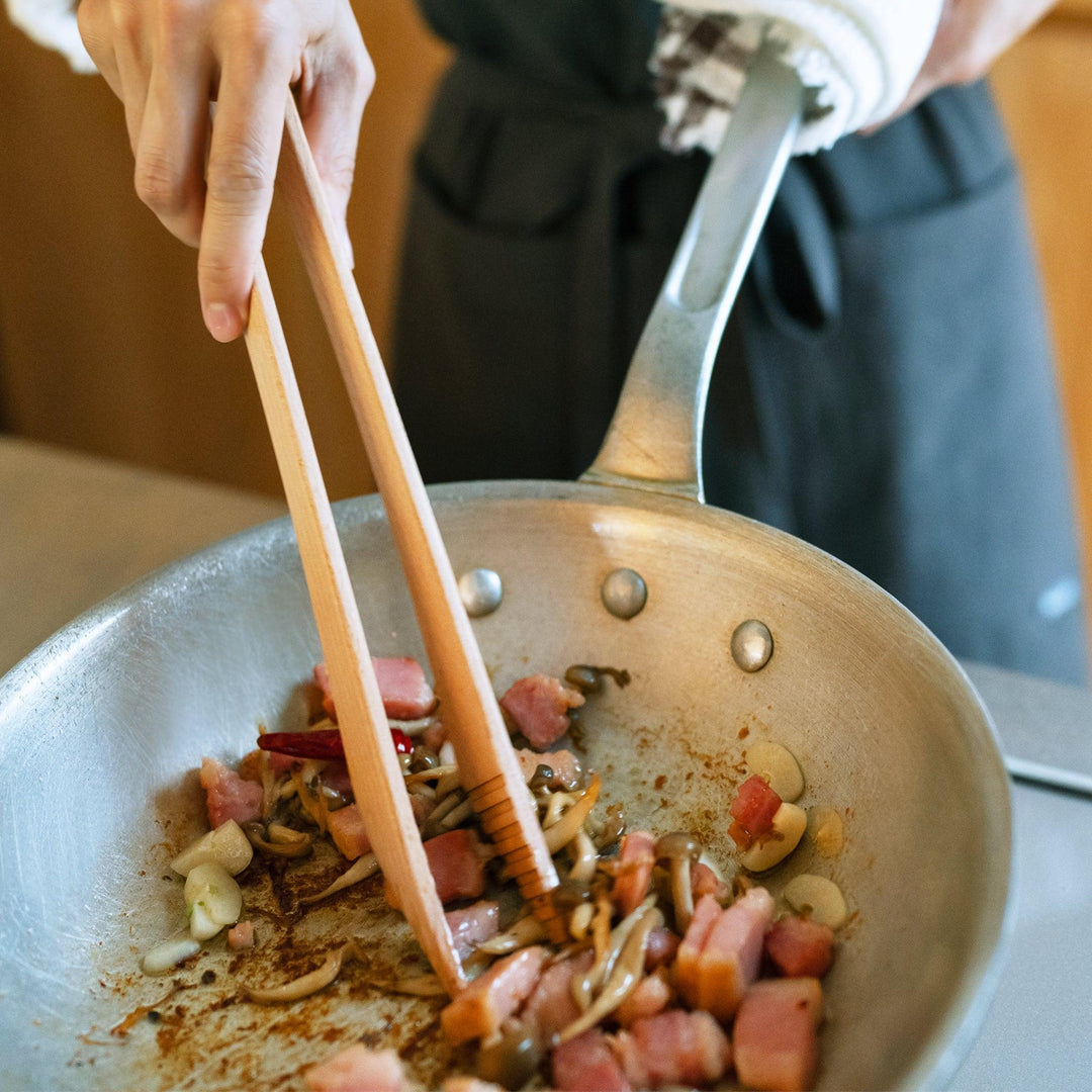 Wooden tongs stirring chopped bacon, mushrooms, garlic, and chili in a frying pan held by a person in a kitchen apron.