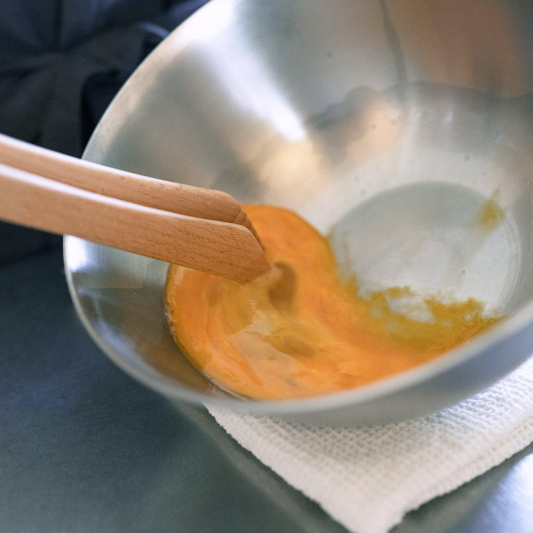 Wooden tongs whisking raw egg in a metal mixing bowl placed on a white cloth on a stainless steel countertop.