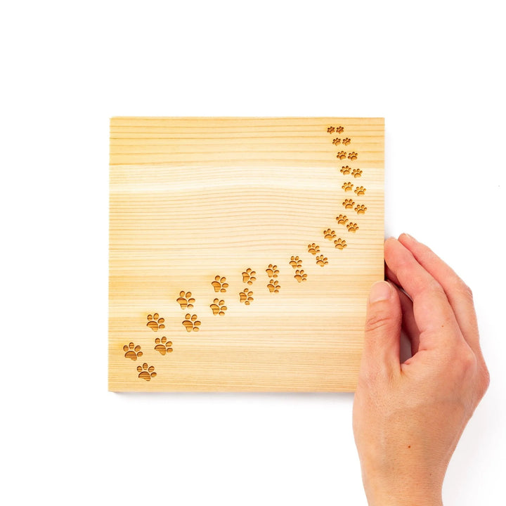 Square hinoki wood trivet featuring curved trail of laser-cut cat paw prints, protecting surfaces with charming feline design