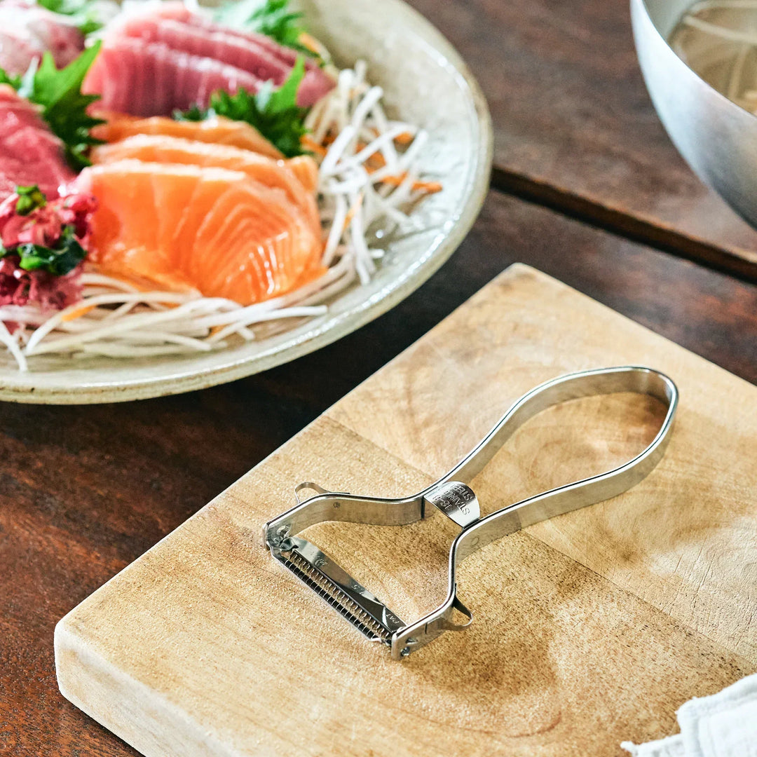A stainless steel julienne peeler rests on a cutting board beside a sashimi platter with daikon garnish.
