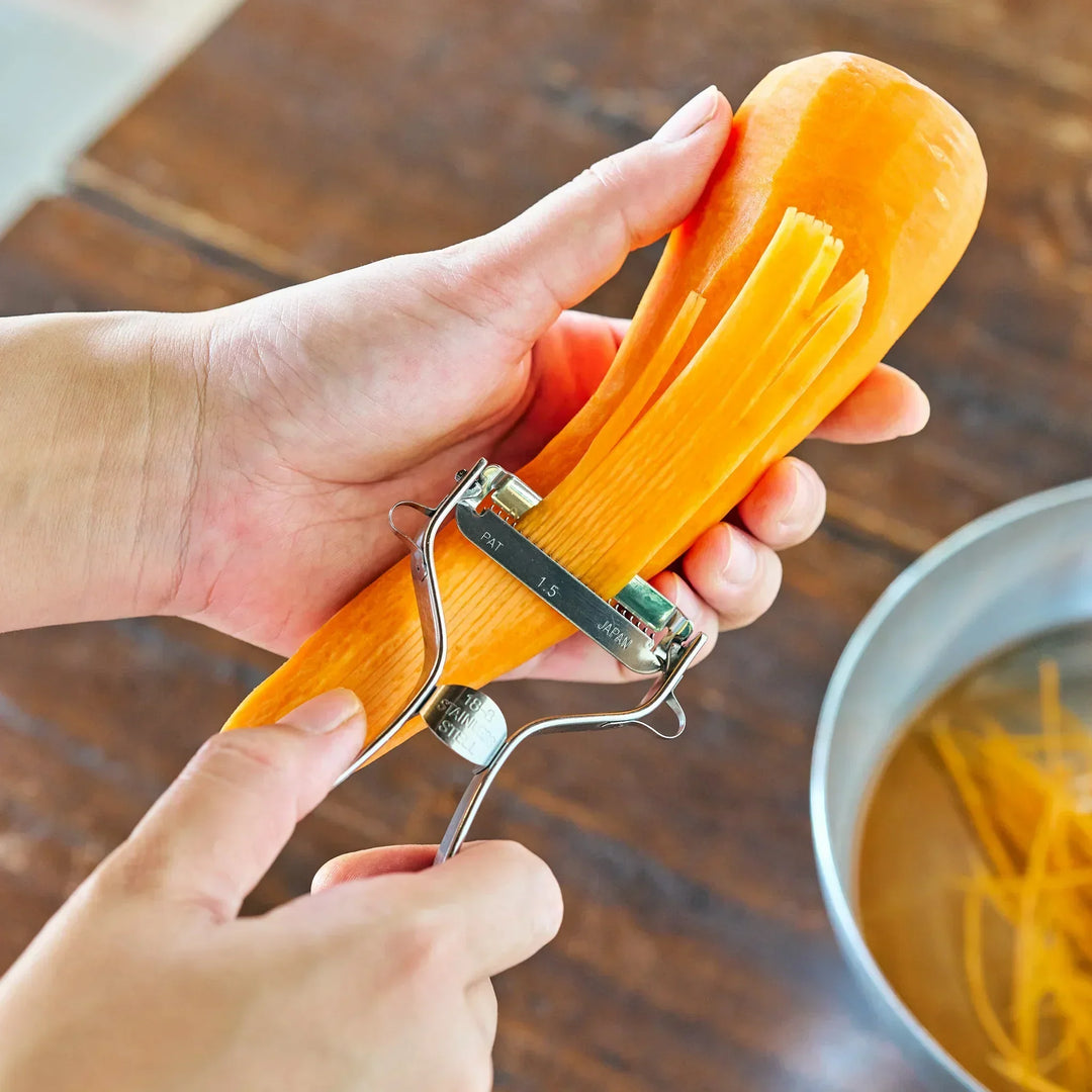 A person shredding a carrot into thin strips with a Japanese julienne peeler over a metal bowl.