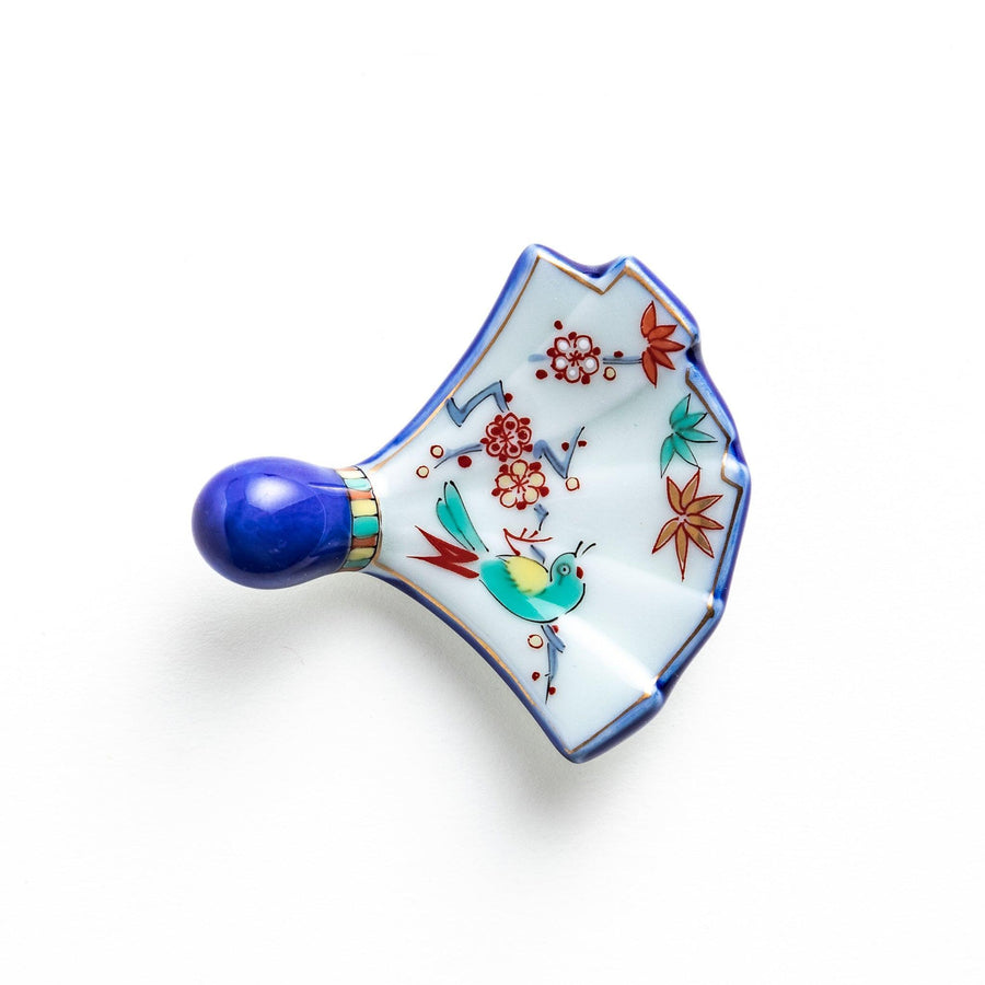 A fan-shaped ceramic chopstick rest decorated with a bird, flowers, and leaves in vibrant colors, with a blue handle.