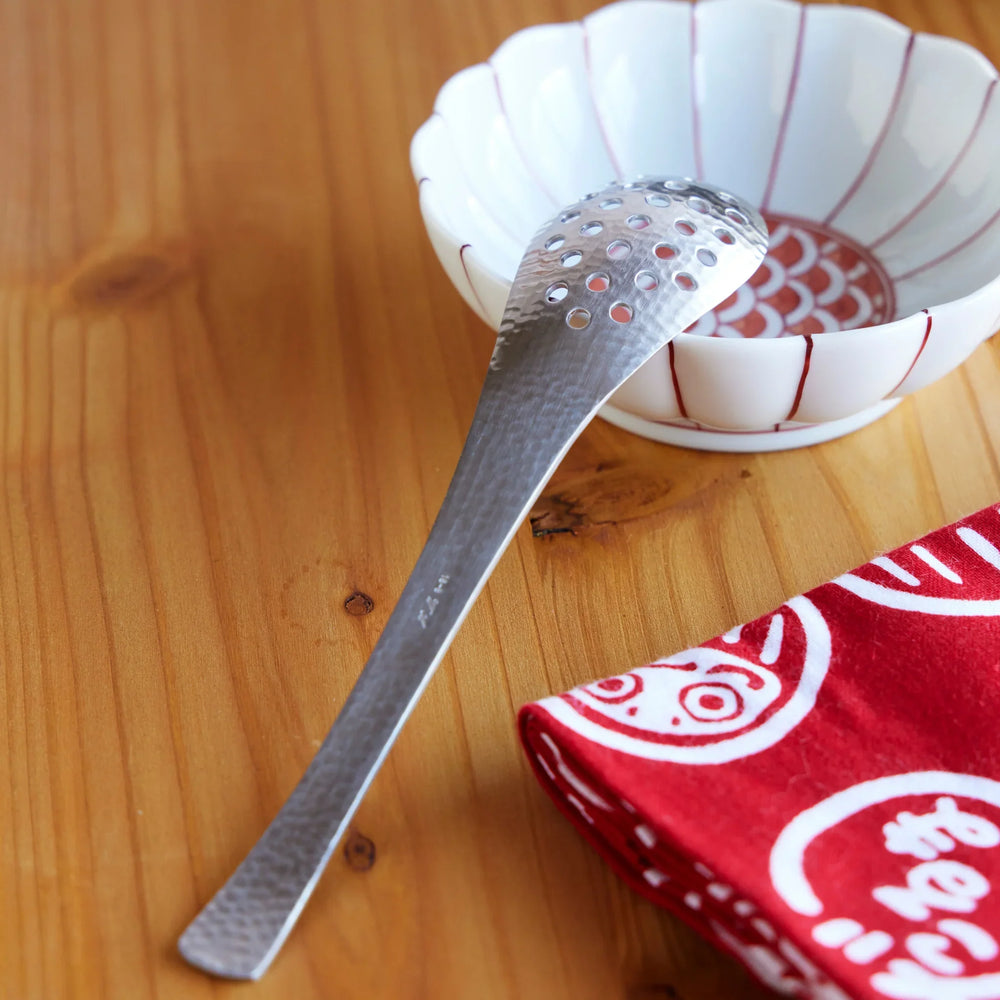 Japanese hammered stainless steel straining spoon with perforated head, resting on porcelain bowl with red pattern design.