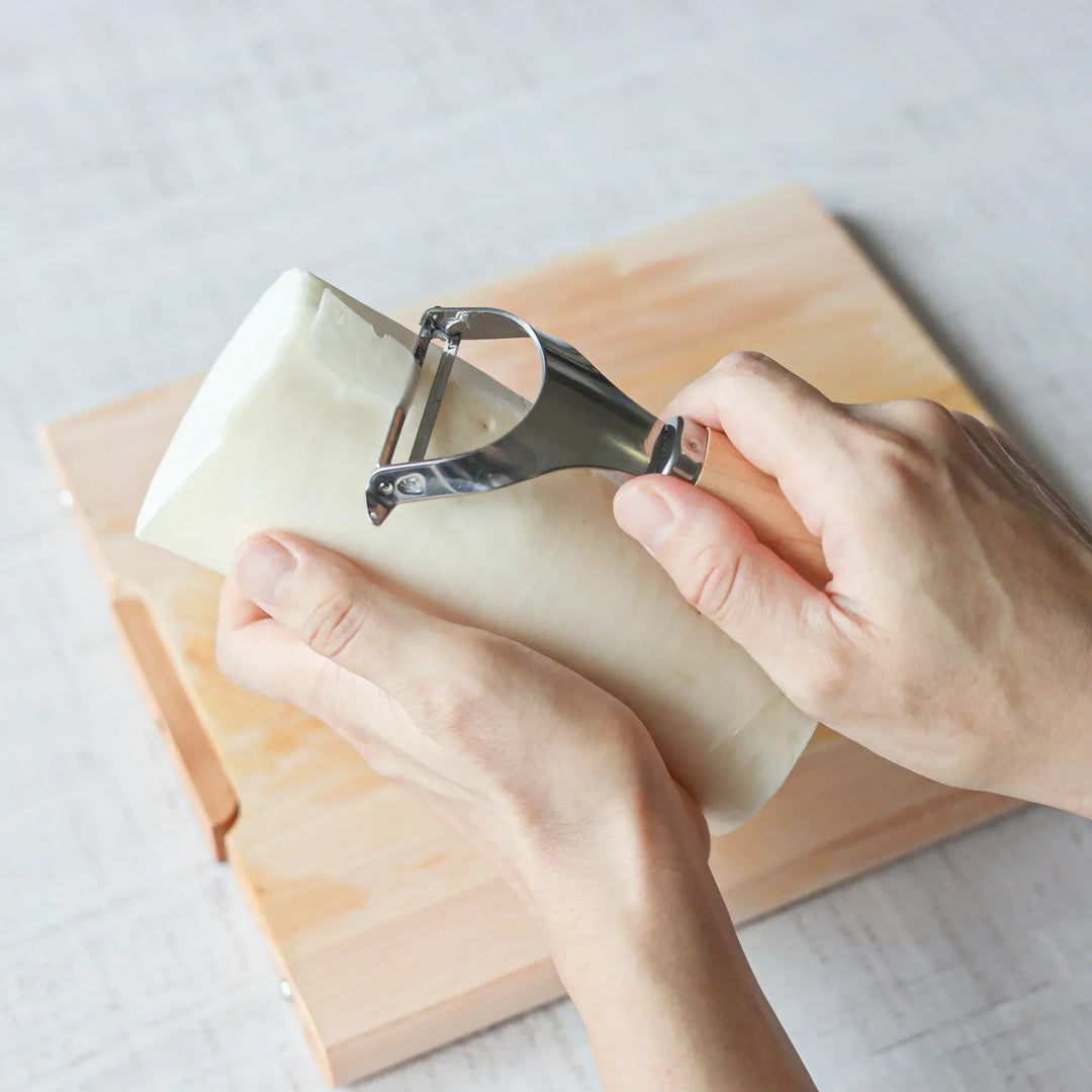 Hands peeling a large daikon radish using a Japanese wood handle stainless steel peeler over a wooden board.
