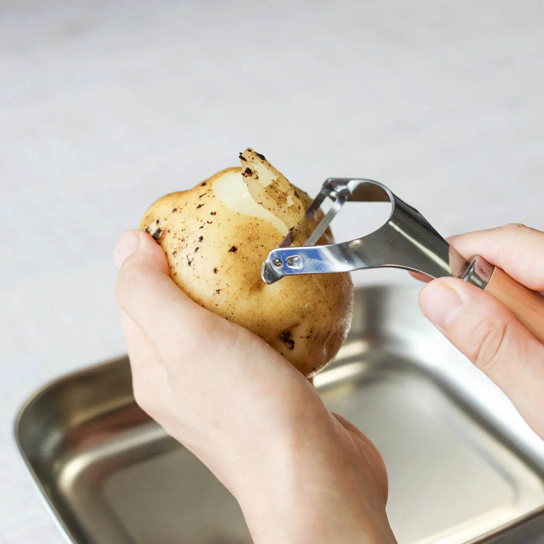 Hands peeling a fresh potato with a Japanese wood handle stainless steel peeler over a metal tray.