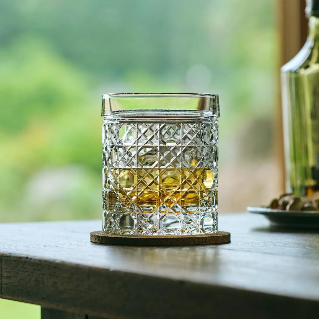 Cut Japanese whiskey glass with a lattice pattern, filled with whisky and ice, on a cork coaster near a window.
