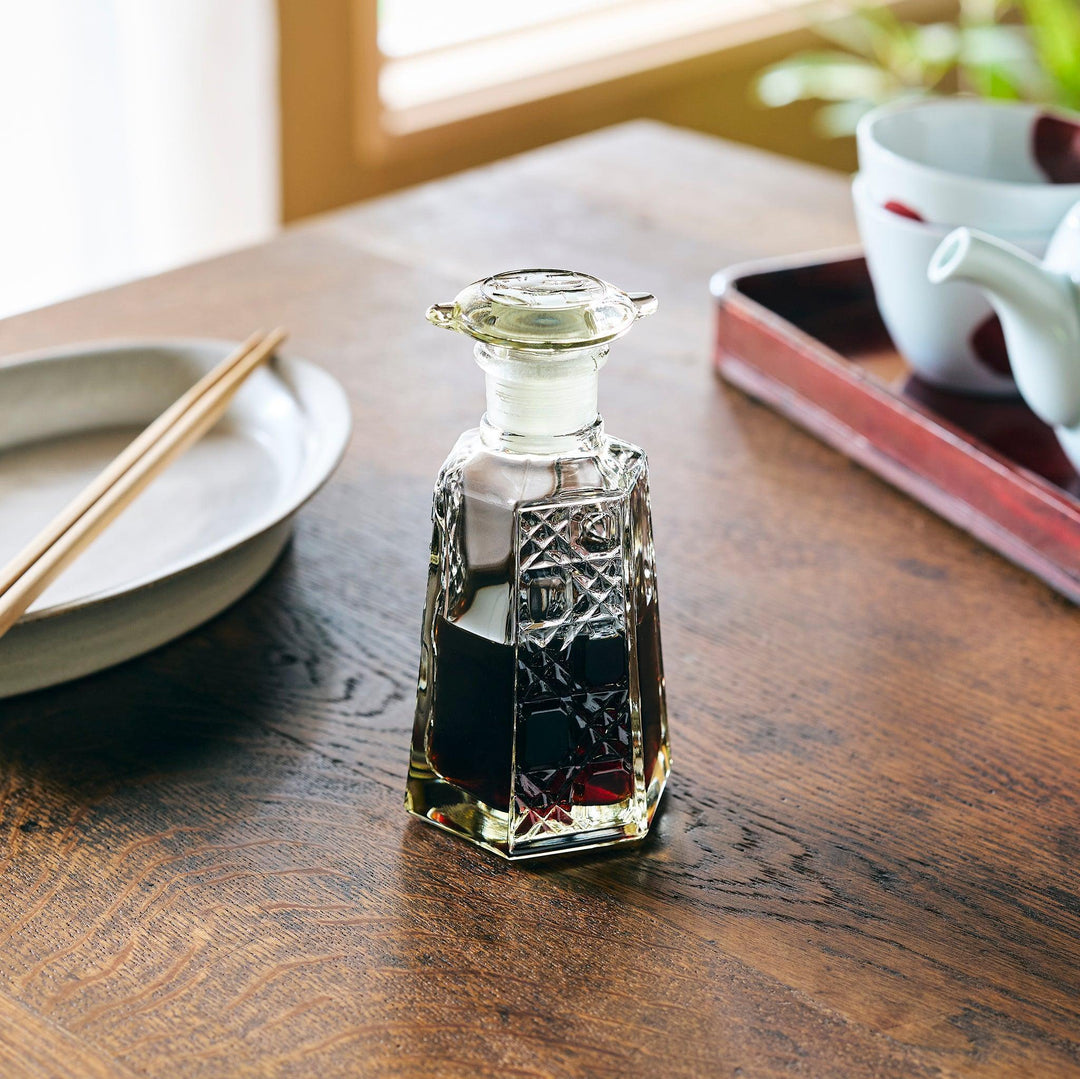Lattice-cut glass soy sauce dispenser with a hexagonal shape, partially filled with dark soy sauce, on a dining table.