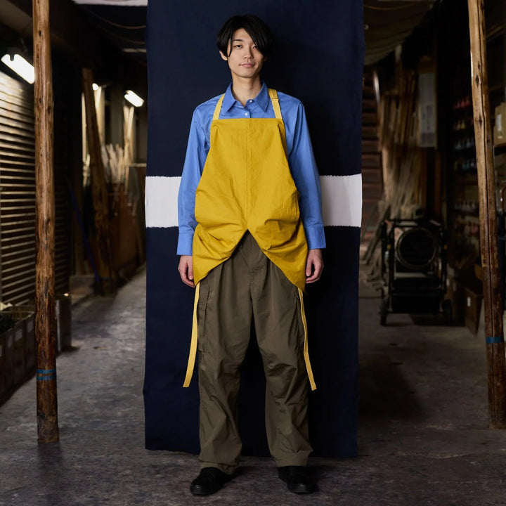 A man wearing a yellow apron with mitt-style ends stands in front of a navy and white backdrop in a rustic workshop.