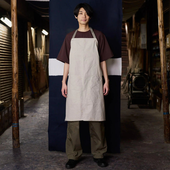 A man wearing a beige apron over a brown shirt and olive pants stands in a rustic workshop with wooden beams and tools.