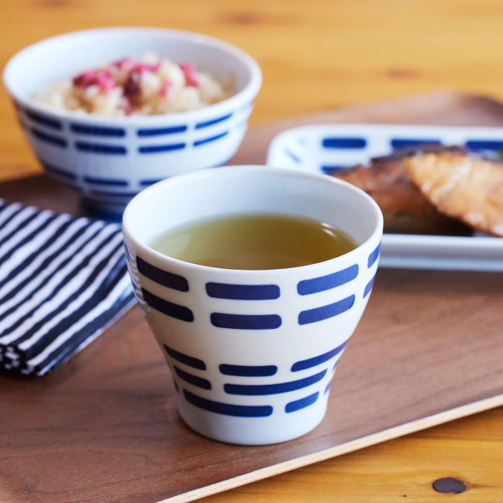 Ceramic cup with indigo horizontal patterns, filled with green tea, placed on a wooden tray with matching tableware