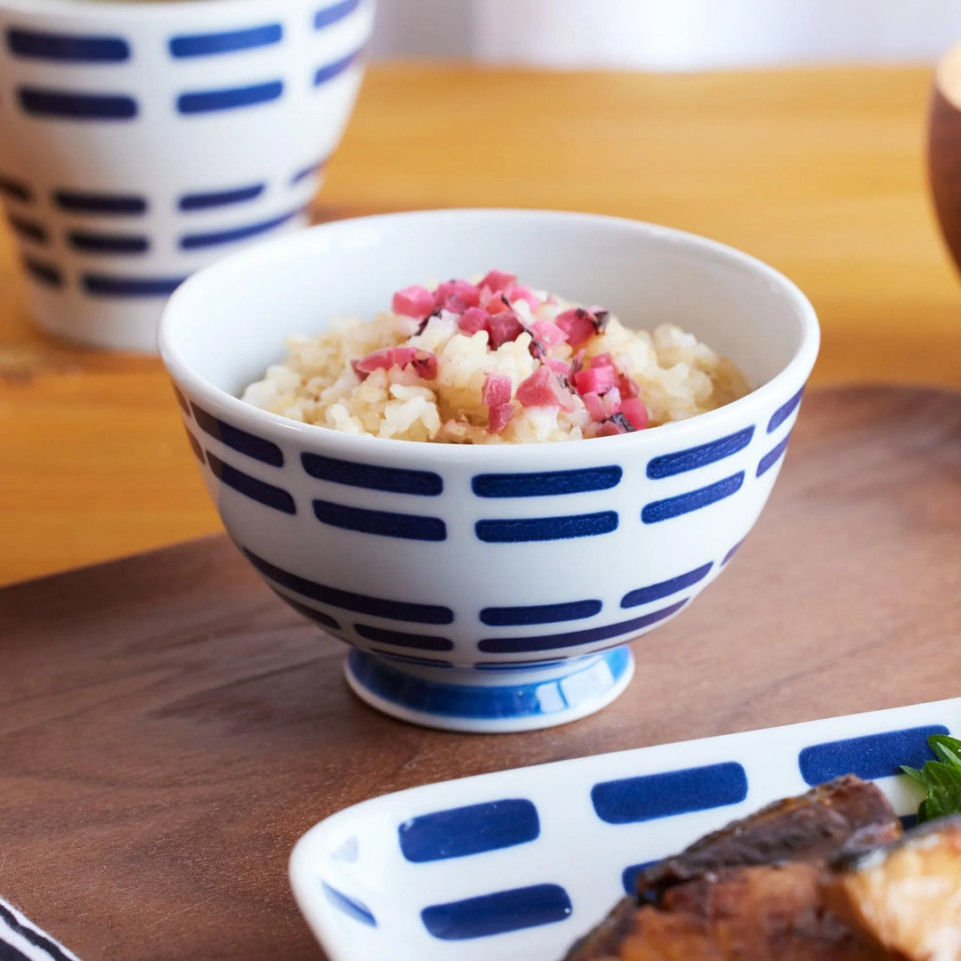 A ceramic rice bowl with indigo horizontal patterns, filled with rice topped with pink pickles, on a wooden tray.